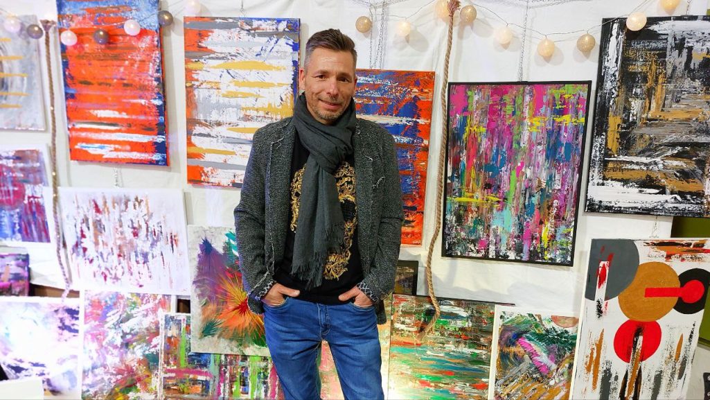 Acryl Galerie & Design Mann in der Mitte vor bunten, abstrakten Kunstwerken an der Wand.