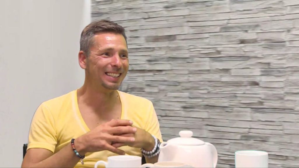 Interview über Vertrieb & Network Marketing Lächelnder Mann in gelbem T-Shirt sitzt am Tisch mit Teekanne und Tassen.